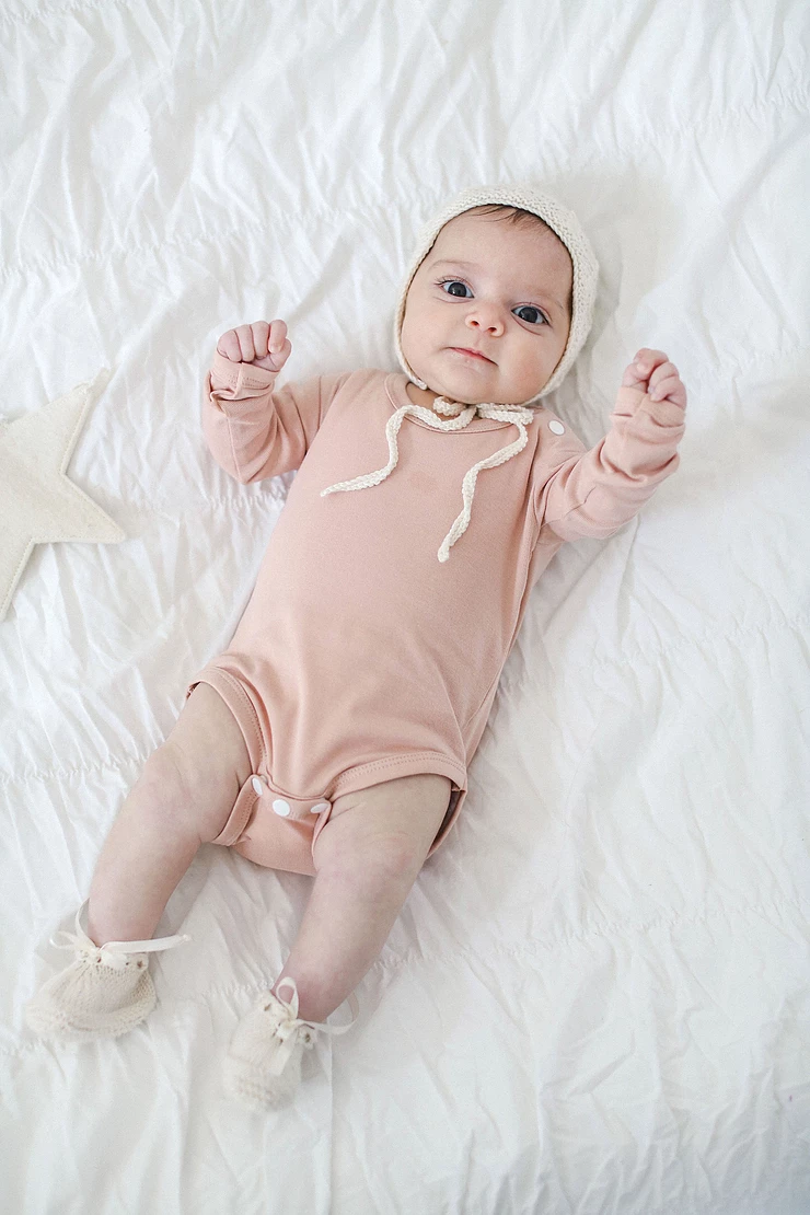 baby girl in soft pink outfit and white knit had, laying on her back on a white blanket
